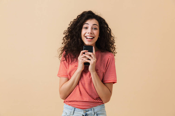 Portrait of an excited young girl with curly hair holding mobile phone isolated over beige background - Photo, Image