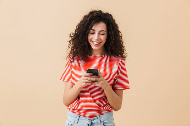 Portrait of a smiling young girl with curly hair using mobile phone isolated over beige background - Φωτογραφία, εικόνα