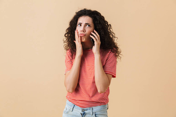 Portrait of a confused young girl with curly hair talking on mobile phone and looking away isolated over beige background - Fotografie, Obrázek
