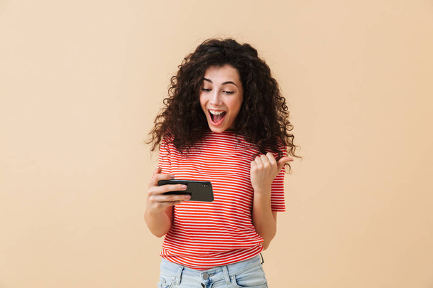 Image of emotional nervous young curly woman standing isolated over beige background play games by mobile phone make winner gesture. - Foto, Bild