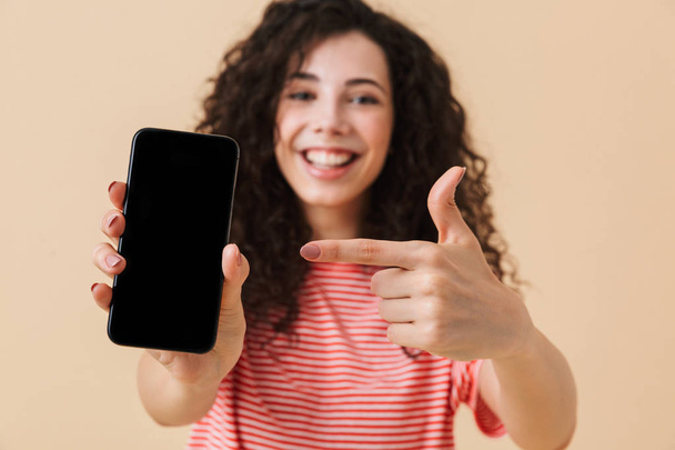 Happy young girl with dark curly hair showing blank screen mobile phone and pointing finger isolated over beige background - Foto, imagen