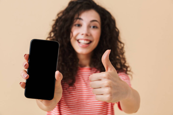 Cheerful young girl with dark curly hair showing blank screen mobile phone and giving thumbs up isolated over beige background - Photo, image