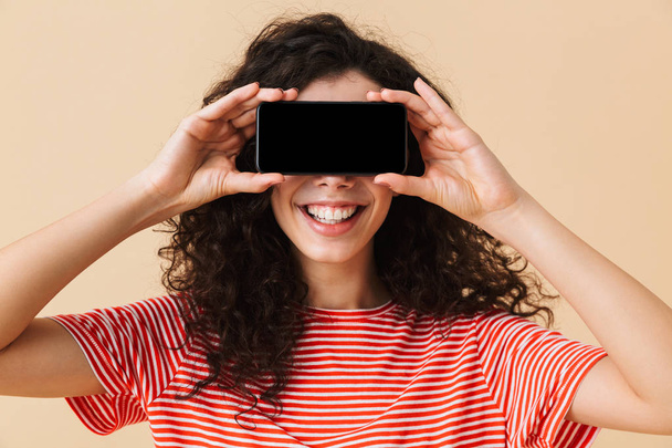 Image of cute young curly woman standing isolated over beige background showing display by mobile phone covering eyes. - Φωτογραφία, εικόνα