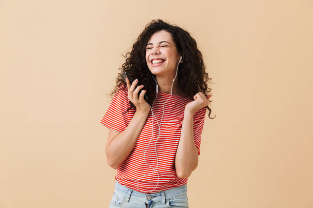 Image of emotional happy excited young curly woman listening music with earphones isolated over beige background dancing. - Φωτογραφία, εικόνα