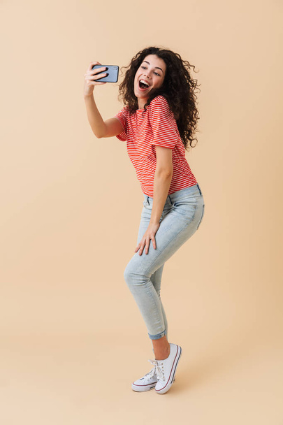 Full length portrait of a cheerful young girl taking a selfie with mobile phone isolated over beige background - Photo, Image