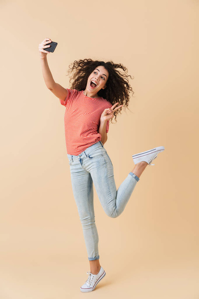 Full length portrait of a joyful young girl taking a selfie with mobile phone while jumping isolated over beige background - Fotó, kép