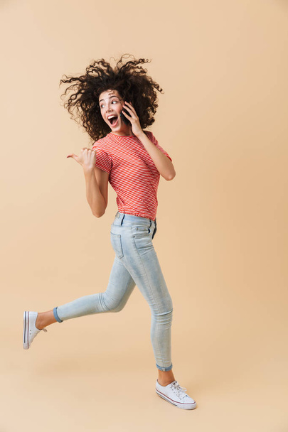 Full length portrait of a cheerful young girl talking on mobile phone while running and pointing away isolated over beige background - Zdjęcie, obraz