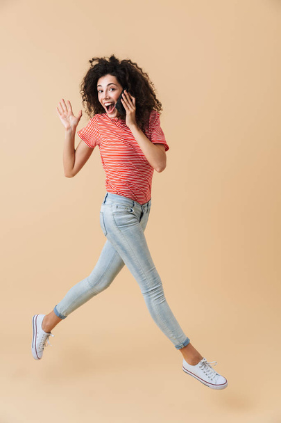 Full length portrait of a cheerful young girl talking on mobile phone while running and waving isolated over beige background - Foto, Imagen