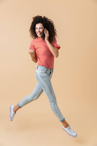 Full length portrait of a cheerful young girl talking on mobile phone while running and celebrating isolated over beige background - Foto, afbeelding