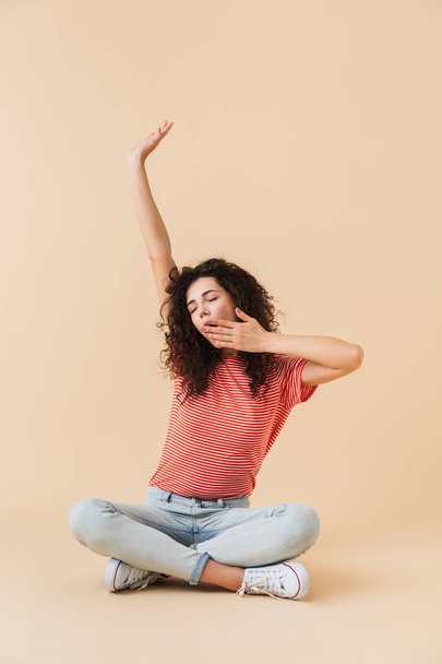 Photo of cute young woman sitting isolated over beige wall background yawning and stretching. - Photo, Image