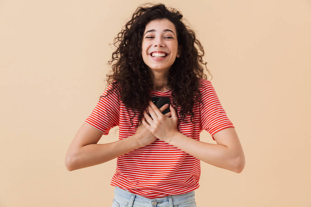 Photo of emotional excited happy young woman isolated over beige wall background using mobile phone. - Foto, Imagem