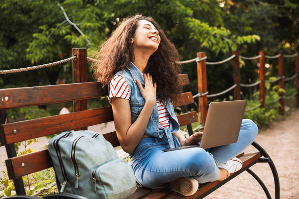 Imagem da mulher relaxada satisfeita com mochila sentada no banco no parque verde no dia de verão e usando laptop prata
 - Foto, Imagem