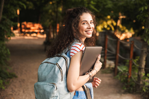 Photo of caucasian young brunette woman 18-20 wearing backpack walking through green park with silver laptop in hands - Фото, зображення