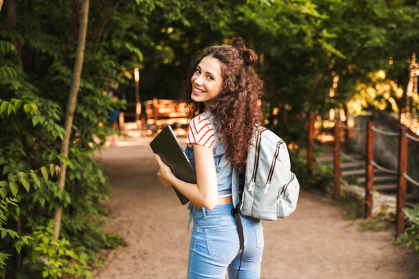 Photo of smiling caucasian woman 18-20 wearing backpack walking through green park with silver laptop in hands - Foto, imagen