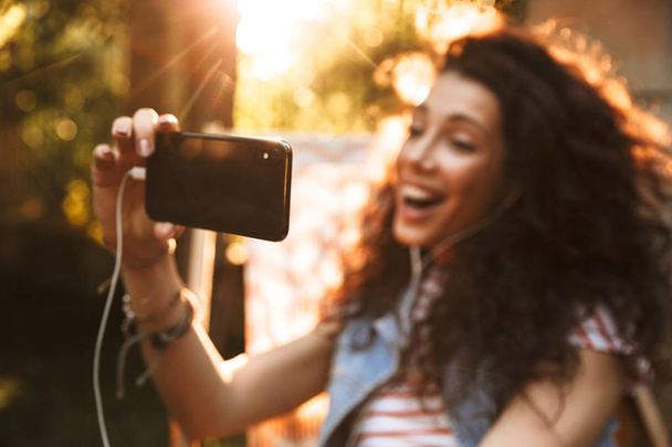 屋外公園でかわいい若い美しい巻き毛女性のイメージはイヤホンで音楽を聴く携帯電話で selfie を作る. - 写真・画像