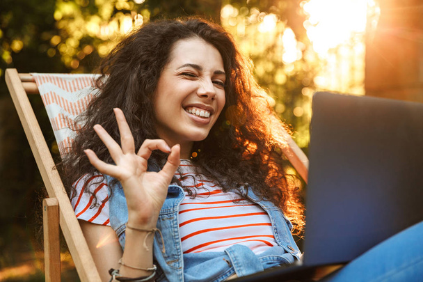 Photo of joyful brunette woman 18-20 showing ok or alright sign while sitting in lounge chair during rest in park on sunny day and using silver laptop - Zdjęcie, obraz