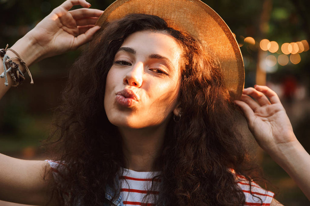 Photo closeup of brunette delighted woman 18-20 wearing summer straw hat blowing air kiss to you while walking through park in sunny day with trees background - Valokuva, kuva