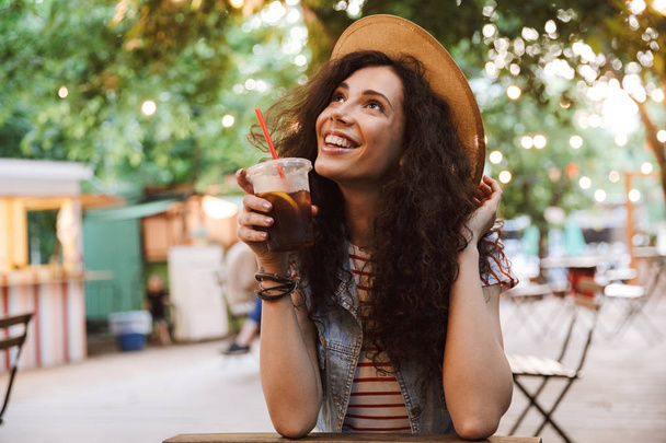 Photo of brunette teenage woman 18-20 wearing summer straw hat drinking cold tea from plastic cup while resting in park cafe - Φωτογραφία, εικόνα