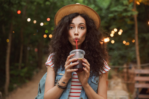Photo of lovely summer woman 18-20 wearing straw hat looking at you while drinking beverage from plastic cup during rest in green park with colorful lamps background - Foto, imagen