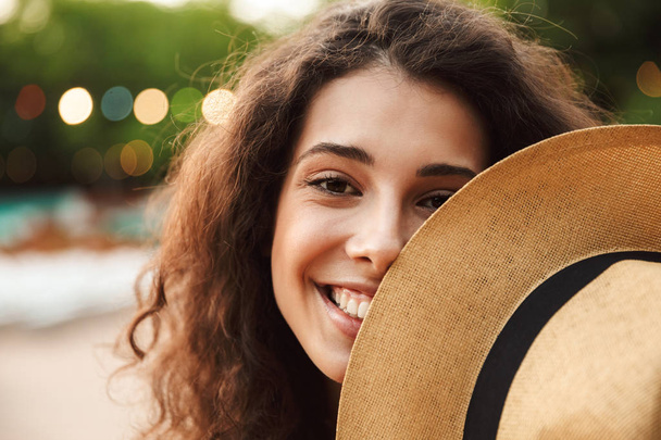 Photo of adorable teen woman 18-20 smiling and covering face with straw hat during walk in green park - Foto, imagen
