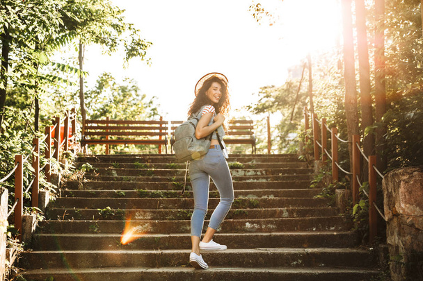 Photo of beautiful teenage girl 18-20 wearing summer clothes and straw hat walking up stairs in green park on sunny day - Foto, imagen