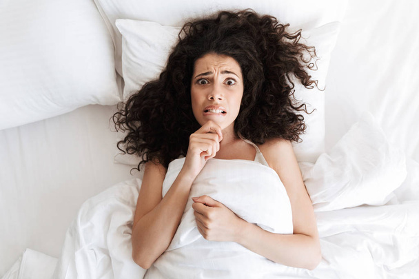 Photo from above of confused young woman 20s with dark curly hair touching chin and lying in white bed with sad emotions - 写真・画像