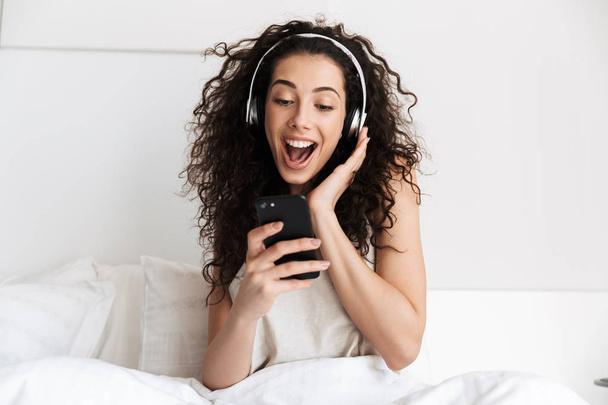 Portrait of cheerful young woman 20s singing while listening to music on smartphone and earphones in white bed at home - Photo, image