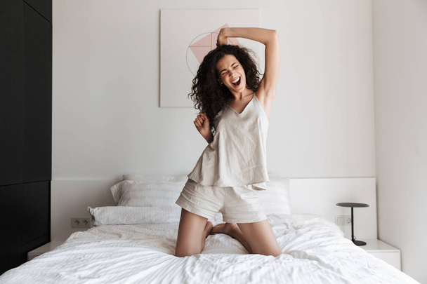 Photo of cheerful woman with long curly hair having fun and dancing while sitting on bed with white clean linen in bedroom - Фото, изображение