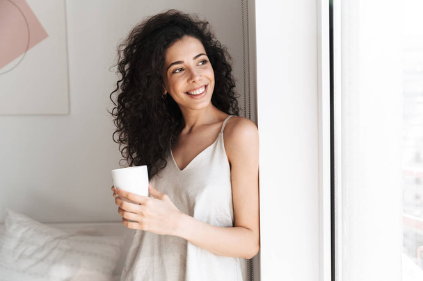 Portrait of gorgeous happy woman with long curly hair looking aside through window in morning at home and holding cup of tea - Zdjęcie, obraz