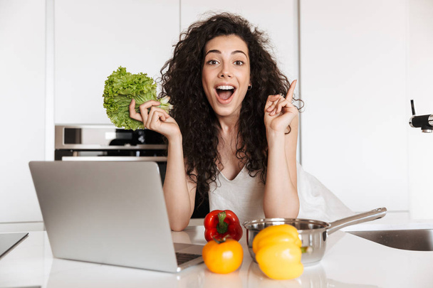 Photo of happy brunette woman 20s holding green salat and reading recipe from laptop while cooking dinner in kitchen interior - Foto, Imagen