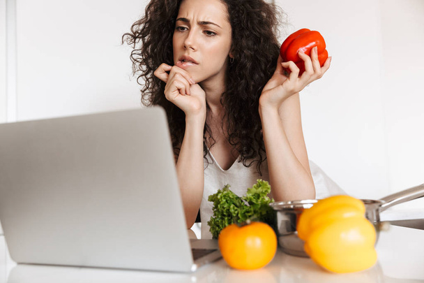 Photo of concentrated curly woman 20s holding sweet paper and looking at screen of laptop while cooking dinner in home kitchen - Фото, изображение