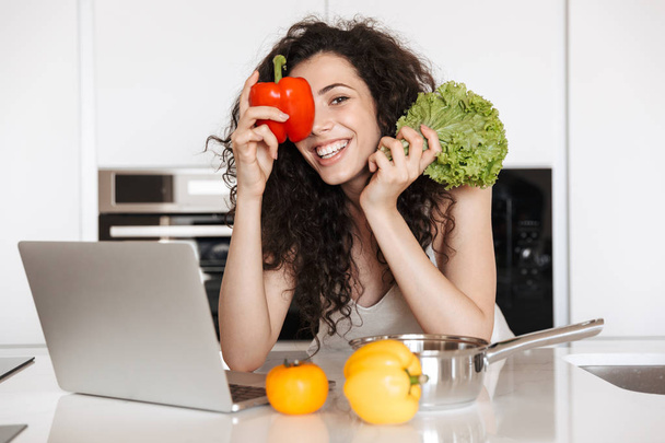 Photo of cheerful woman vegetables lover holding green salat and sweet paper while cooking dinner in kitchen and reading recipe from laptop - Foto, afbeelding
