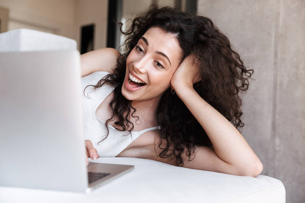 Photo closeup of young adorable woman with long curly hair wearing silk leisure clothing lying on couch in apartment and laughing while looking at screen of laptop - Foto, Bild