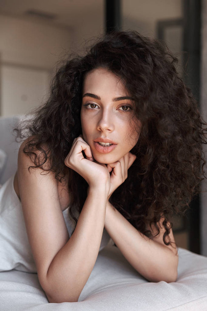 Caucasian adorable woman with long curly hair wearing silk leisure clothing lying on couch in apartment, and looking at you while propping up head with hands - Photo, Image