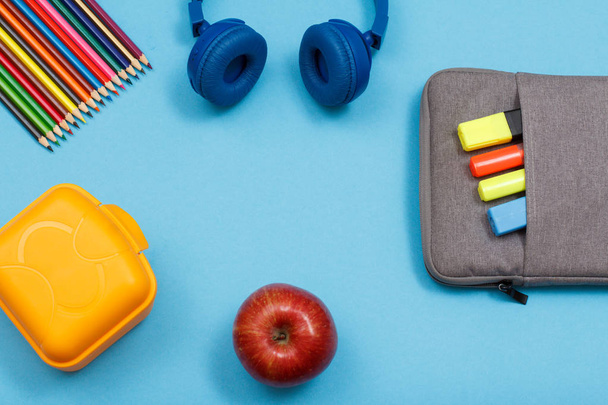 Lunch box, color pencils, headphones, apple and bag-pencil case with color felt pens and marker on blue background. Top view with copy space. Back to school concept. School supplies - Фото, зображення