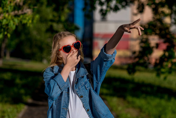 Girl schoolgirl. Summer in nature. Sunglasses in the shape of the heart. Fingers his hand points to side. The concept of surprise surprise. Emotion of delight indignation. - Фото, изображение