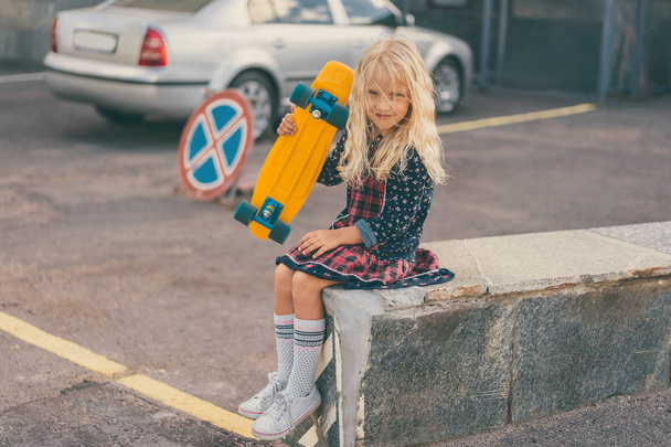 adorable smiling kid holding skateboard and sitting at urban street  - Фото, зображення