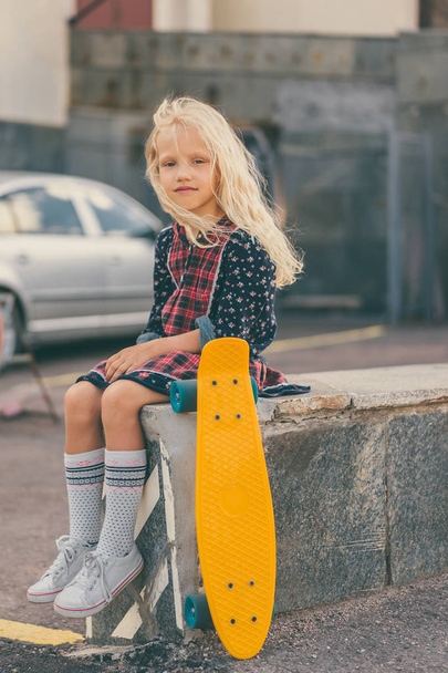 adorable child sitting near skateboard and looking at camera at urban street - Foto, afbeelding
