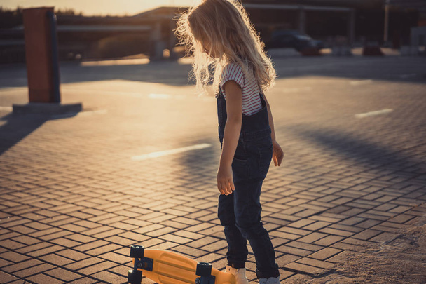 Zijaanzicht van klein kind permanent in de buurt van skateboard op parkeerplaats  - Foto, afbeelding