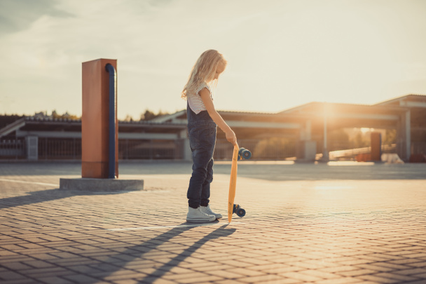 vista lateral del niño pequeño de pie con penny board en el estacionamiento
 - Foto, imagen