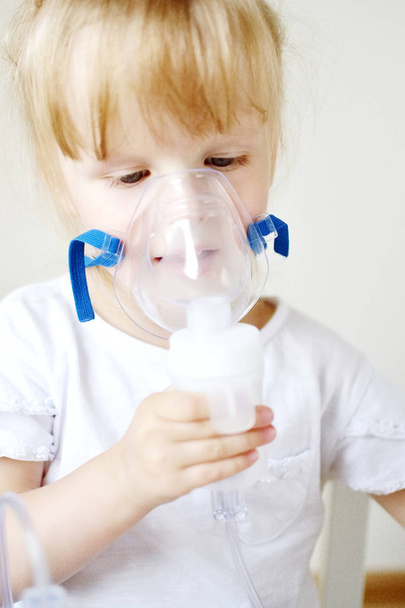 Little girl in a mask for inhalations, making inhalation with nebulizer at home inhaler on the table, indoor, sick child - Photo, image