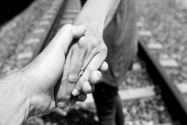 closeup of the hand of a caucasian man holding the hand of another caucasian man, seen from behind, walking by the railroad tracks, in black and white - Photo, Image