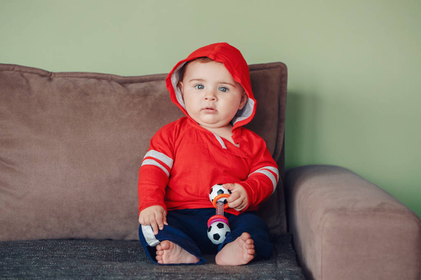 Portrét roztomilý rozkošný kavkazských chlapeček, nosí červené sportovní mikina tričko a tepláky kalhoty. Sedm měsíců dítě sedí na gauči a držení fotbal fotbal báchorka - Fotografie, Obrázek