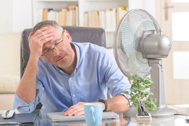 Чоловік страждає від спеки під час роботи в офісі і намагається охолодити вентилятор
 - Фото, зображення