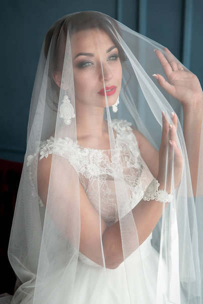 Red lips elegant, young brunette bride in vintage white dress posing under veil closeup with red lips. Bride portrait wedding makeup and hairstyle, fashion bride. Beautiful bride in veil - Photo, Image