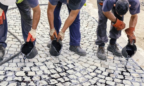 Teamwork on pouring bitumen or thick tar into the joints. Traditional pitch jointing for paved driveway. Using pitch bucket, spreading liquid tar to apply the joint of road. Pitch jointing for cubes. - Photo, Image