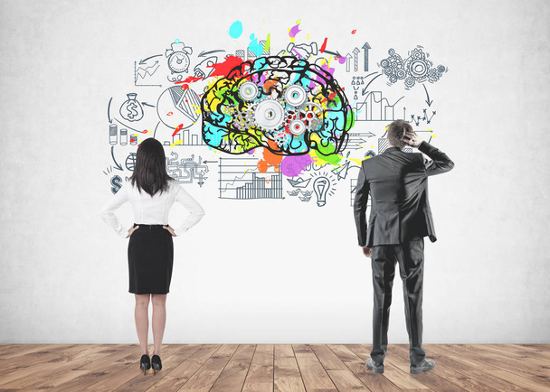 Rear view of a businesswoman with long black hair and a businessman in a gray suit scratching his head and looking at a concrete wall with a cog brain and business scheme sketches on it. - Foto, Imagen