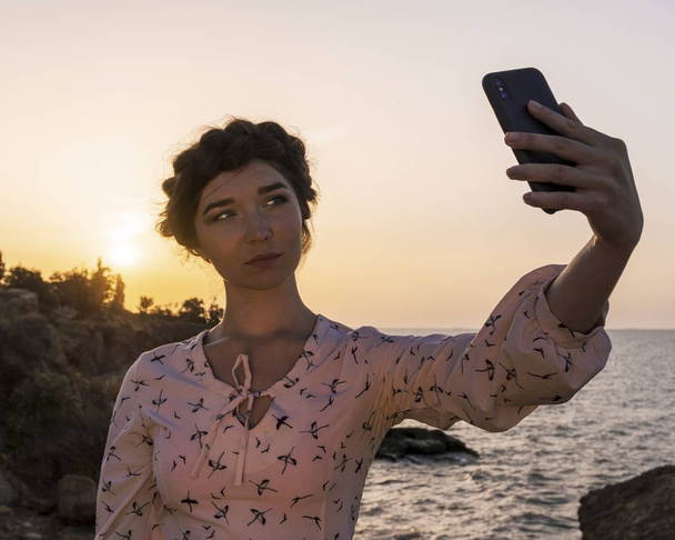 Αξιολάτρευτο κορίτσι Μελαχρινή καυκάσιος στο φως φόρεμα απολαμβάνοντας με το τηλέφωνο στο Seacoast Sunrise στιγμή. - Φωτογραφία, εικόνα