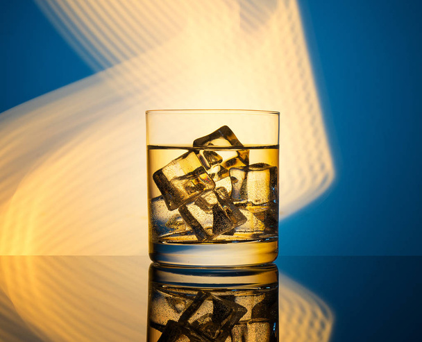 A glass with a drink and ice cubes on a blue background with golden lights. - Foto, imagen