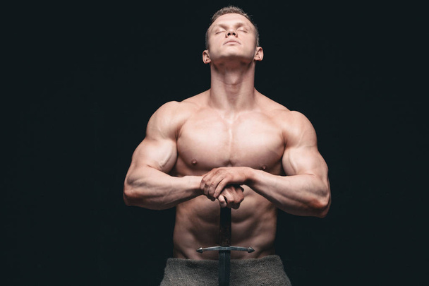 Bodybuilder man posing with a sword isolated on black background. Serious shirtless man demonstrating his mascular body. hands on a sword - Foto, Imagem